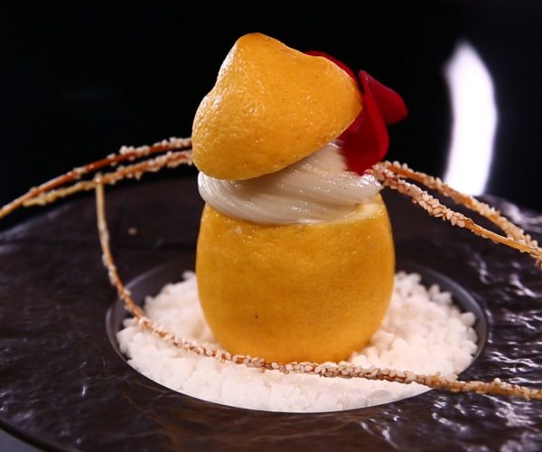 Dessert au citron recréé en forme de fruit, servi sur un lit de sucre et décoré de fins fils de caramel et d'une fleur comestible